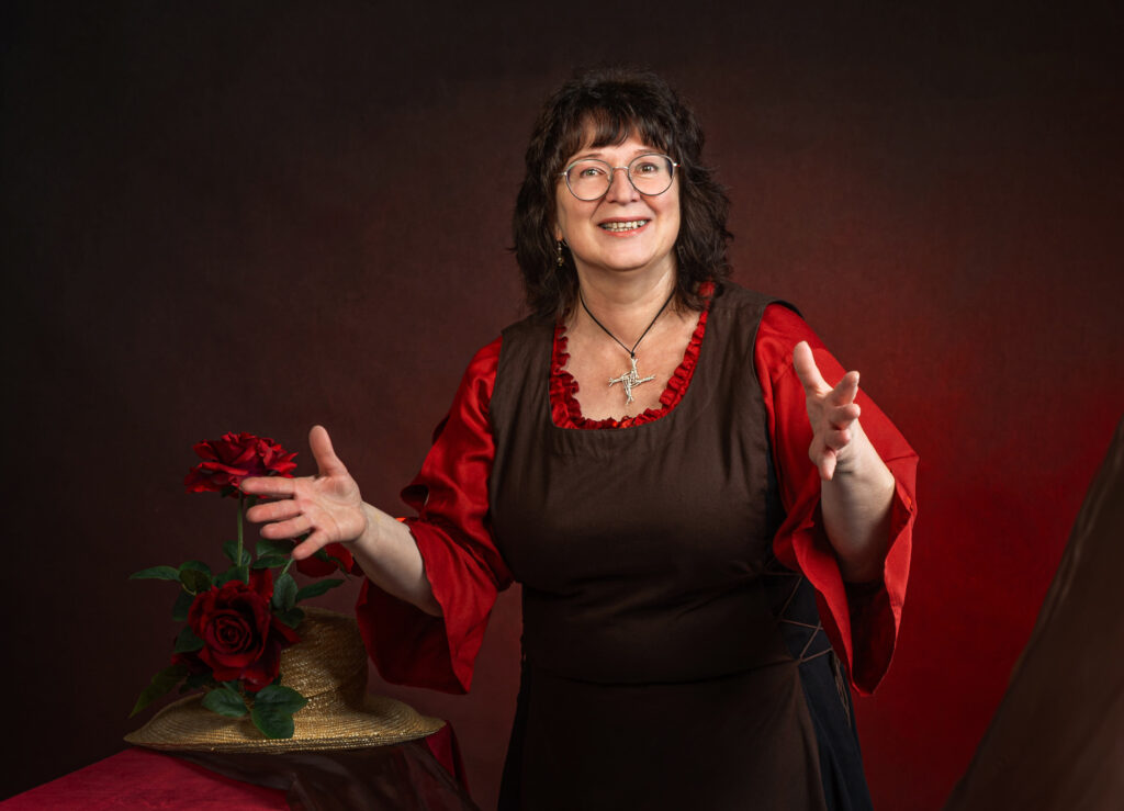 Erzählerin Christine Graf im mittelalterlichen Kleid mit Rosenhut; lächelnd in erzählender Geste vor rot-braunem Hintergrund.